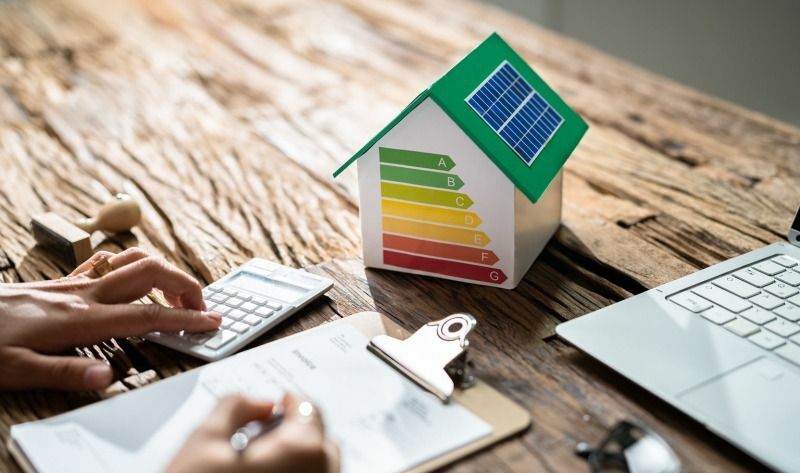 Hands using calculator alongside a clipboard, with a model house featuring the energy rating scale on its side.