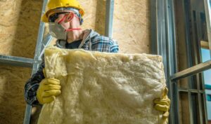 Contractor wearing protective gear and carrying a batt of fiberglass insulation in a new building construction site.