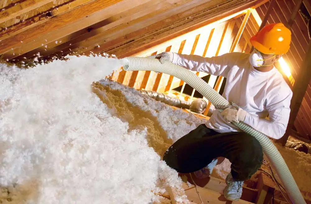 Worker spraying fiberglass insulation in attic, wearing a hardhat and mask.