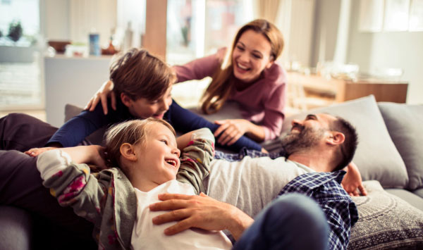 Happy family of four (mom, dad, and two kids) laughing and playing together on couch.