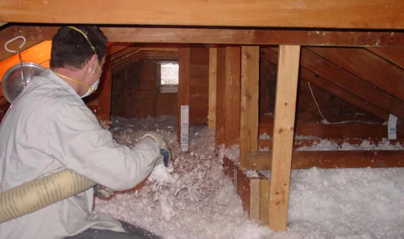 Technician in attic spraying blown-in white insulation between wooden beams
