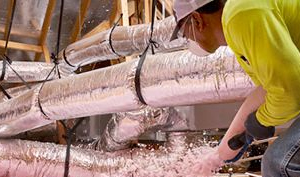 Technician in yellow shirt installing pink fiberglass insulation around foil-wrapped HVAC ducts in attic