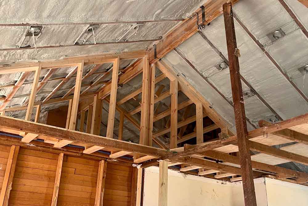 An interior view of a building under construction, showing a vaulted ceiling insulated with white spray foam between wooden rafters.