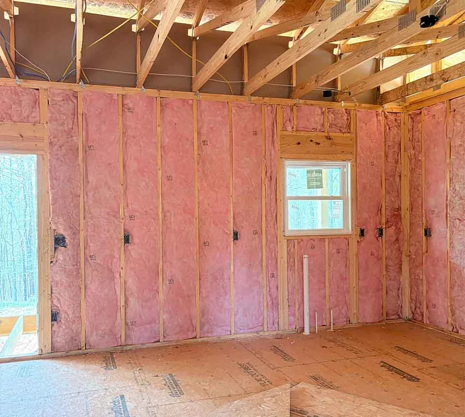 Pink fiberglass batt insulation installed in framed walls of a new construction home with open roof trusses above.