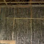 Unfaced fiberglass batt insulation filling wood-framed wall cavities in a garage with exposed roof trusses.
